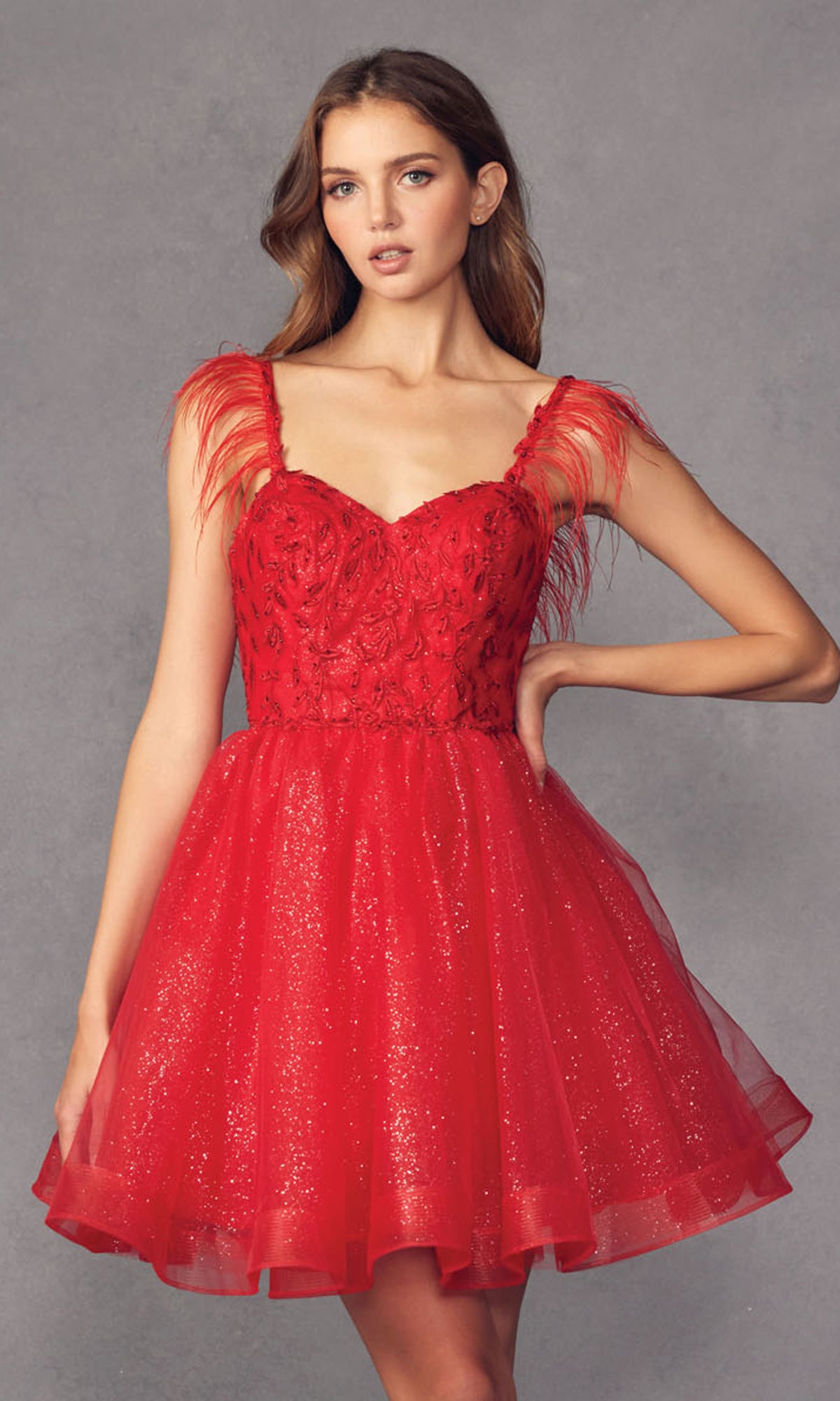 A woman poses against a plain gray background, wearing the Short Babydoll Homecoming Dress with Feathers 881—a glittery dress featuring feathered shoulder straps, a sparkly tulle skirt, and an embroidered sweetheart bodice.